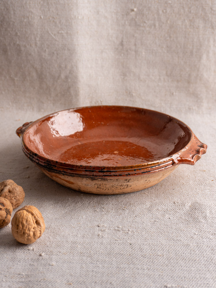 Mid-century Vallauris terracotta baking dish with handles on textured beige fabric, three walnuts beside it.