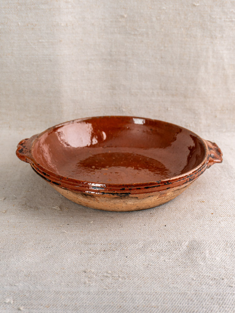 Mid-century Vallauris terracotta baking dish with handles, glossy interior, rustic exterior on light textured fabric.