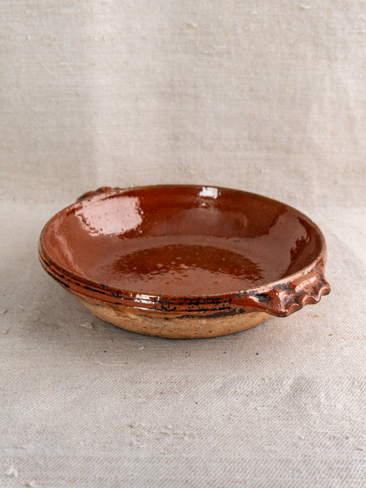 Mid-century Vallauris terracotta baking dish with handles and glossy brown glaze on textured beige fabric.