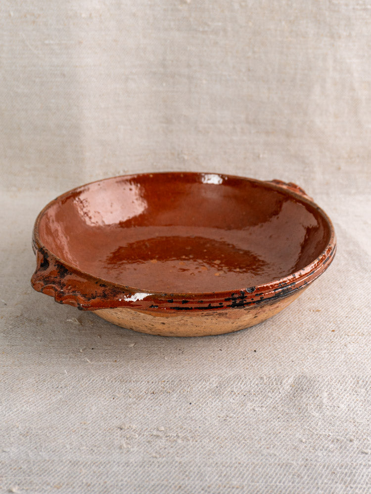 Mid-century Vallauris terracotta baking dish with glossy finish and handles on textured beige fabric backdrop.