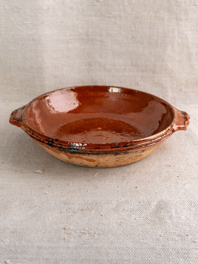 Mid-century Vallauris terracotta baking dish with handles and brown glaze on beige textured fabric background.