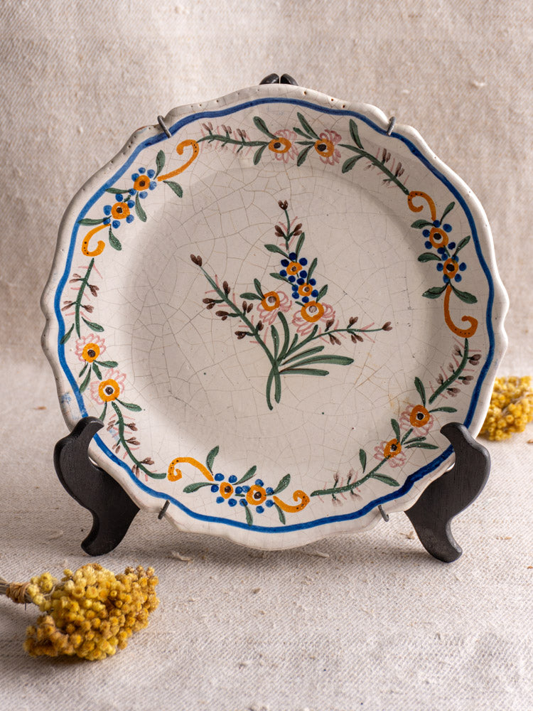 Antique French faience plate with hand-painted blue, orange, and green floral motifs on a black stand with dried yellow flowers.