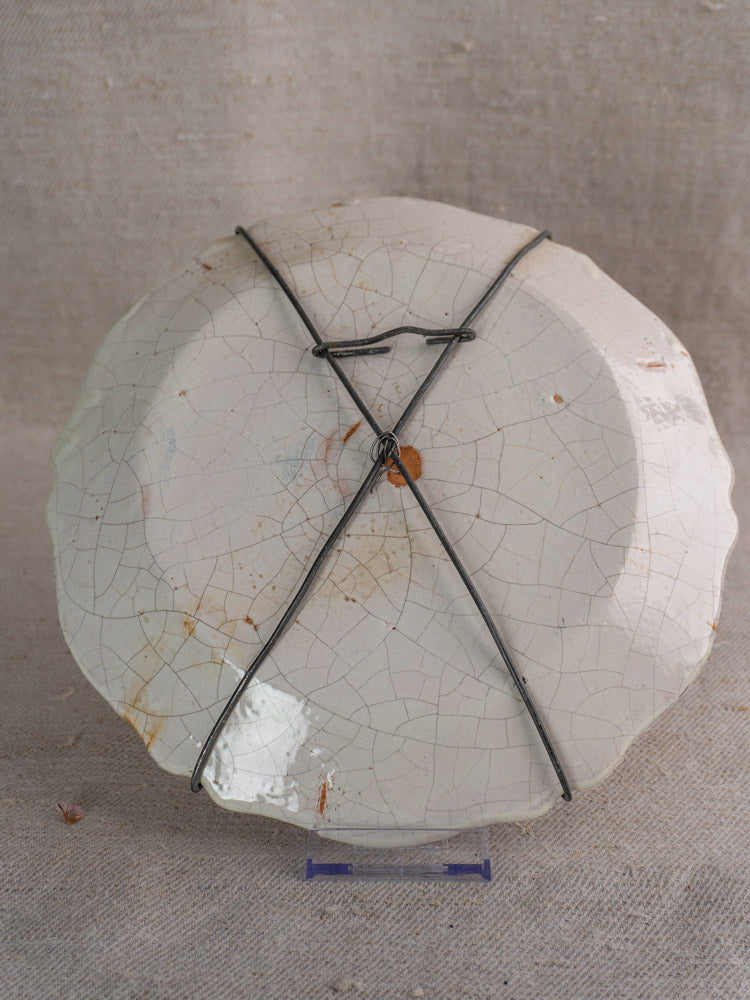 Back of antique French faience plate with stains, black metal wire hanger, on neutral fabric background.