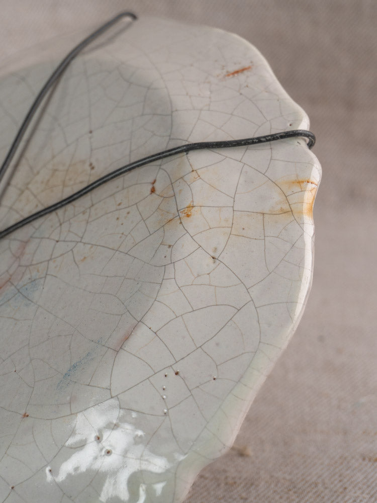 Antique French faience plate with hand-painted florals, pale cracked glaze, subtle stains, and wire across ceramic on fabric background.