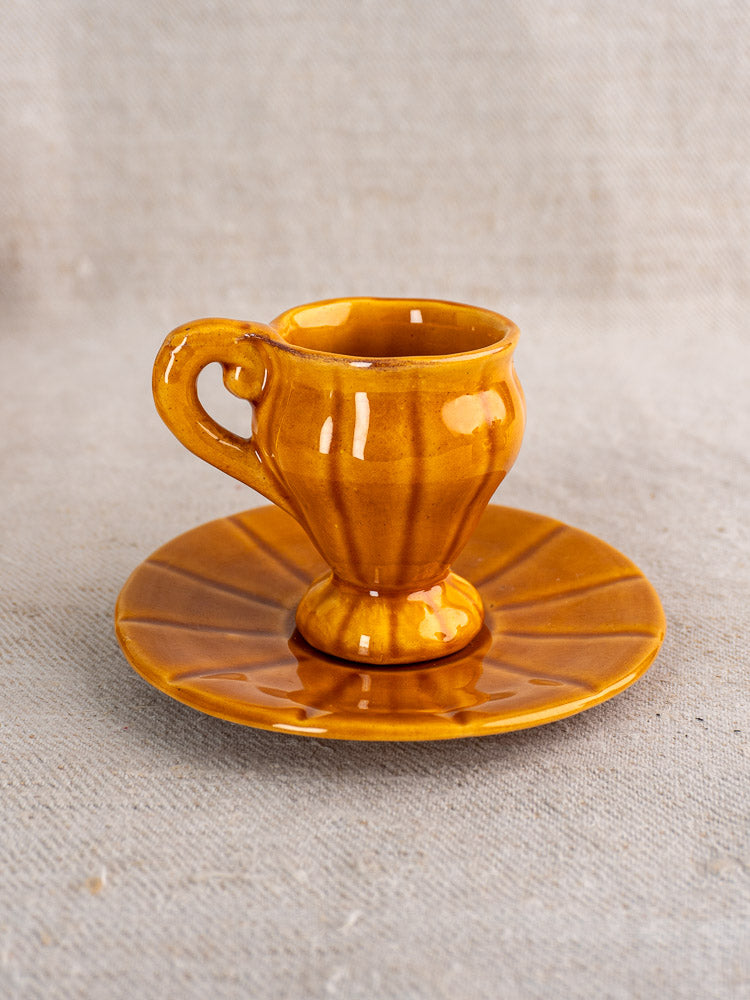 Vintage French honey-glazed earthenware demitasse cup and saucer with ridged pattern on fabric surface.