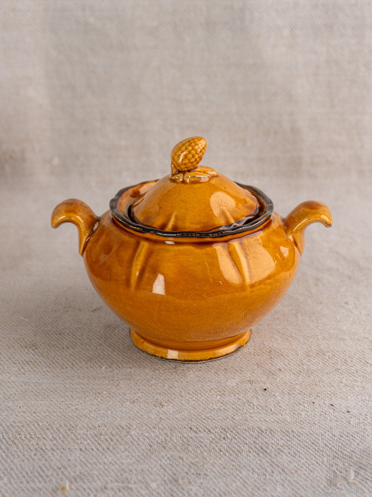 Honey-glazed earthenware sugar bowl with two handles, acorn lid, on textured beige fabric.