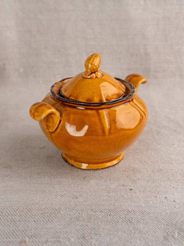 Small round brown ceramic demitasse pot with honey-glazed finish and decorative knob on beige fabric.