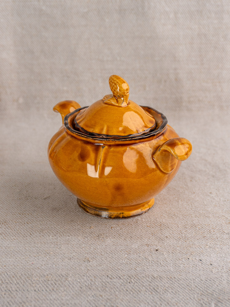 Small round brown ceramic teapot with lid and dual handles on textured beige fabric background.
