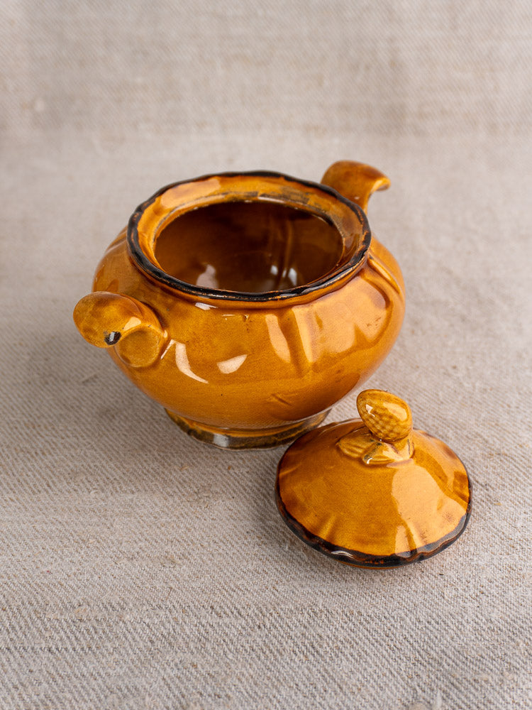 Small round honey-glazed earthenware pot with two handles and lid on textured fabric.