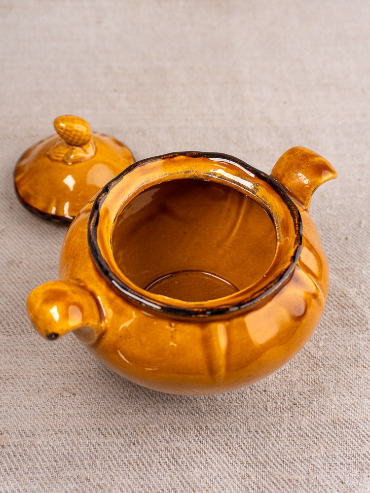 Small round brown glazed earthenware pot with side handles, matching lid beside on beige fabric.