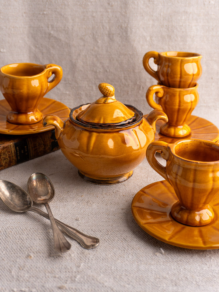 Vintage French honey glazed earthenware demitasse coffee set with teacups, saucers, teapot, and silver spoons on textured cloth.