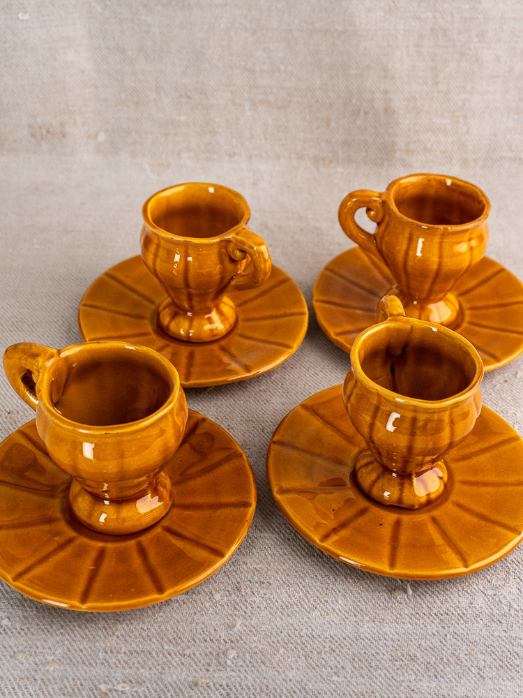Set of four honey-glazed earthenware demitasse cups with saucers, featuring rounded handles and subtle radial-patterned saucers on a beige fabric surface.