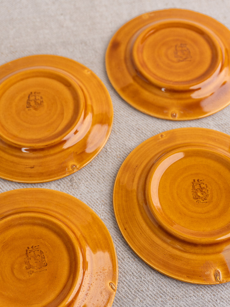 Four honey-glazed earthenware demitasse plates displayed upside down, showcasing maker’s marks on beige textured surface.
