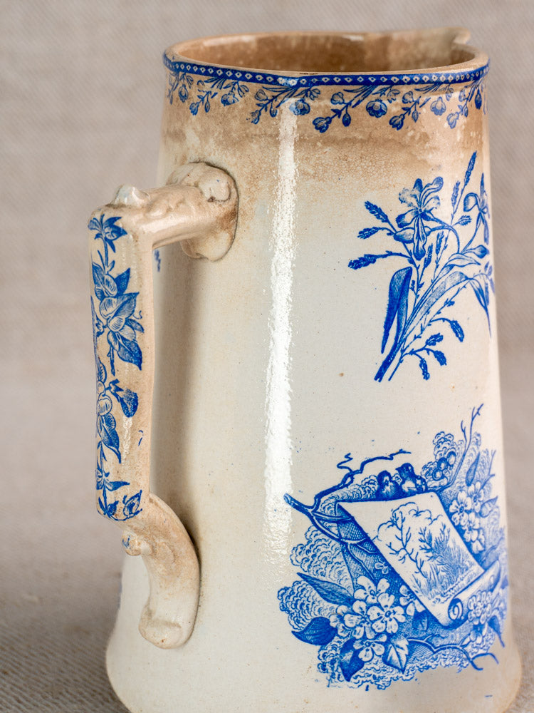 Antique French Fenal Frères ceramic pichet in Pexonne Oiseaux pattern with blue floral motifs, brown-cream gradient, on fabric background.