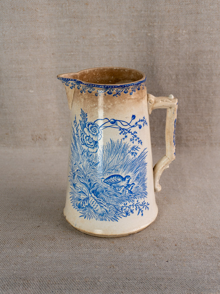 Antique French Fenal Frères Pichet with blue floral motif and ornate handle on beige fabric.