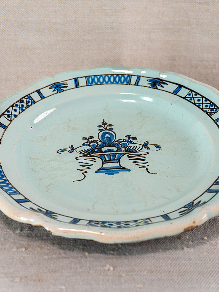 19th-century French faience plate with scalloped edge, blue floral center, Rouen geometric rim, on beige textured surface.