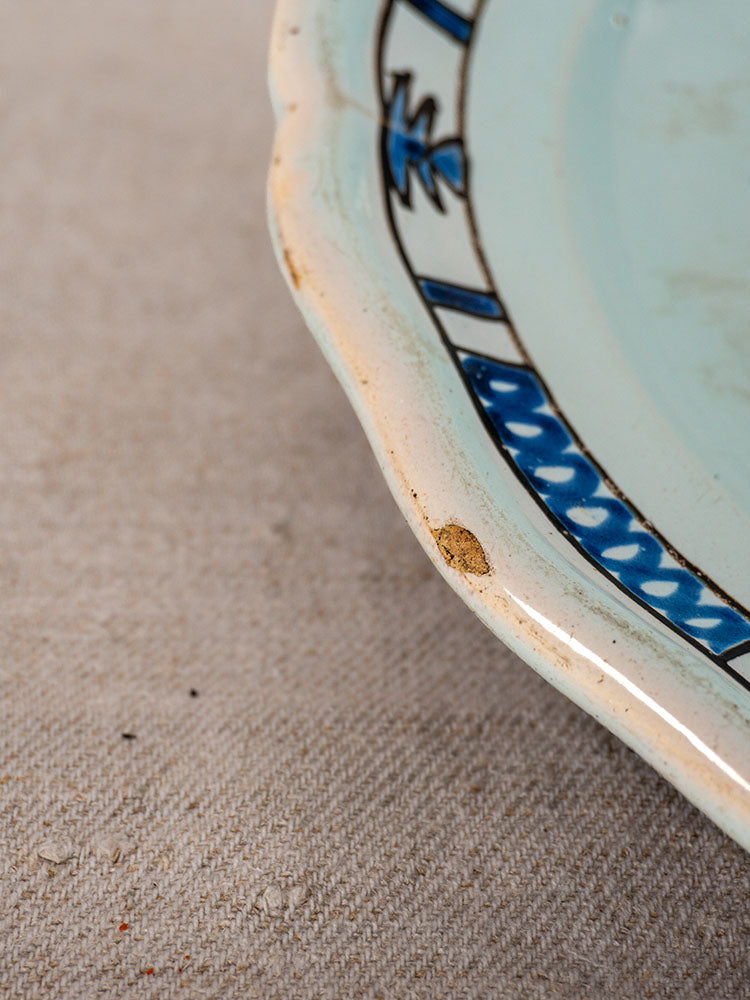 Antique French faience plate with blue and black border, small rim chip, shown on textured beige fabric.