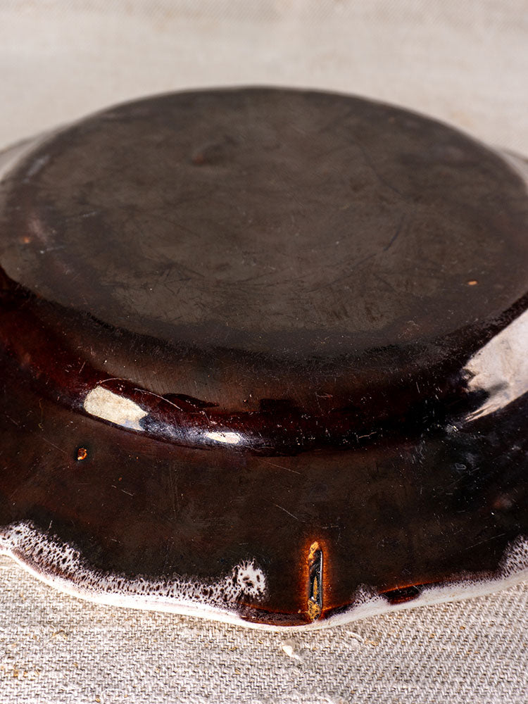 Antique French faience plate, Cul Noir, upside down on textured fabric; rim chips expose reddish layer under dark brown glaze.