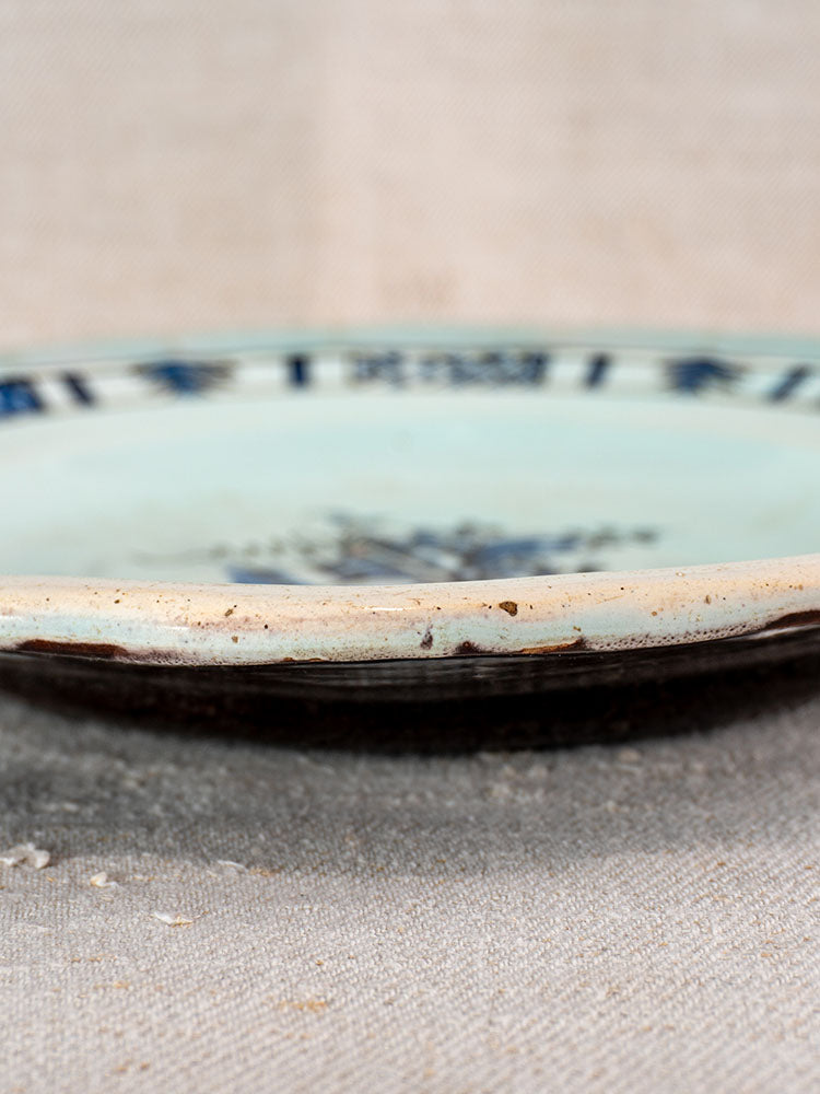Antique French faience plate with light blue glaze, dark blue decor, and rim wear on beige textured fabric.