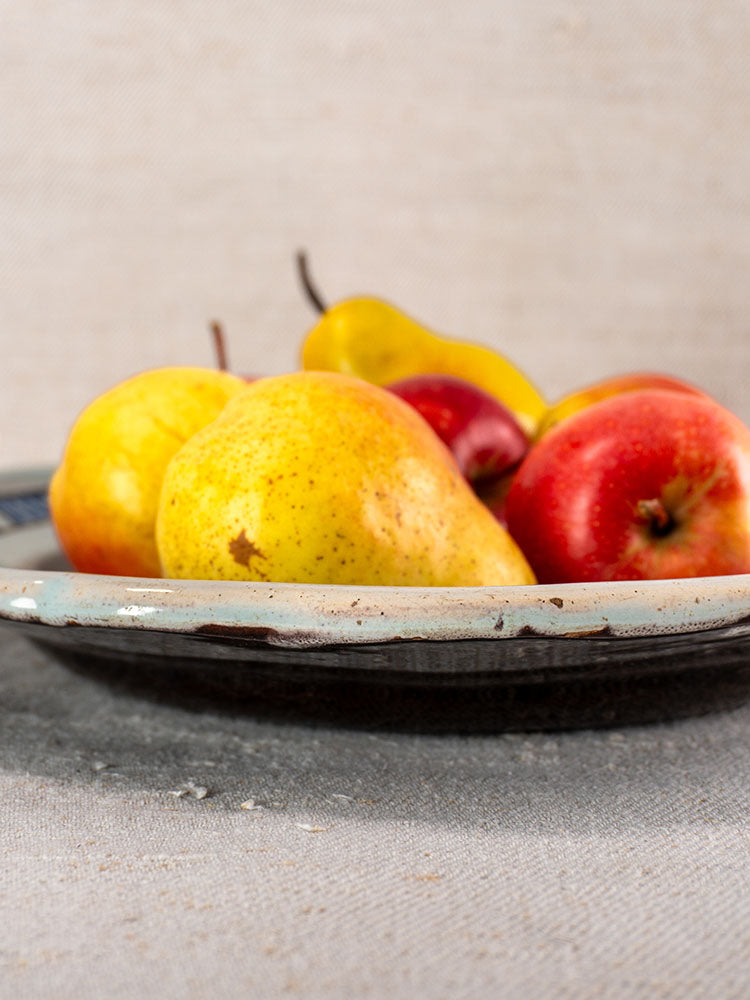 Antique French faience Cul Noir plate with yellow pears and red apples on light gray textured surface, neutral fabric backdrop.