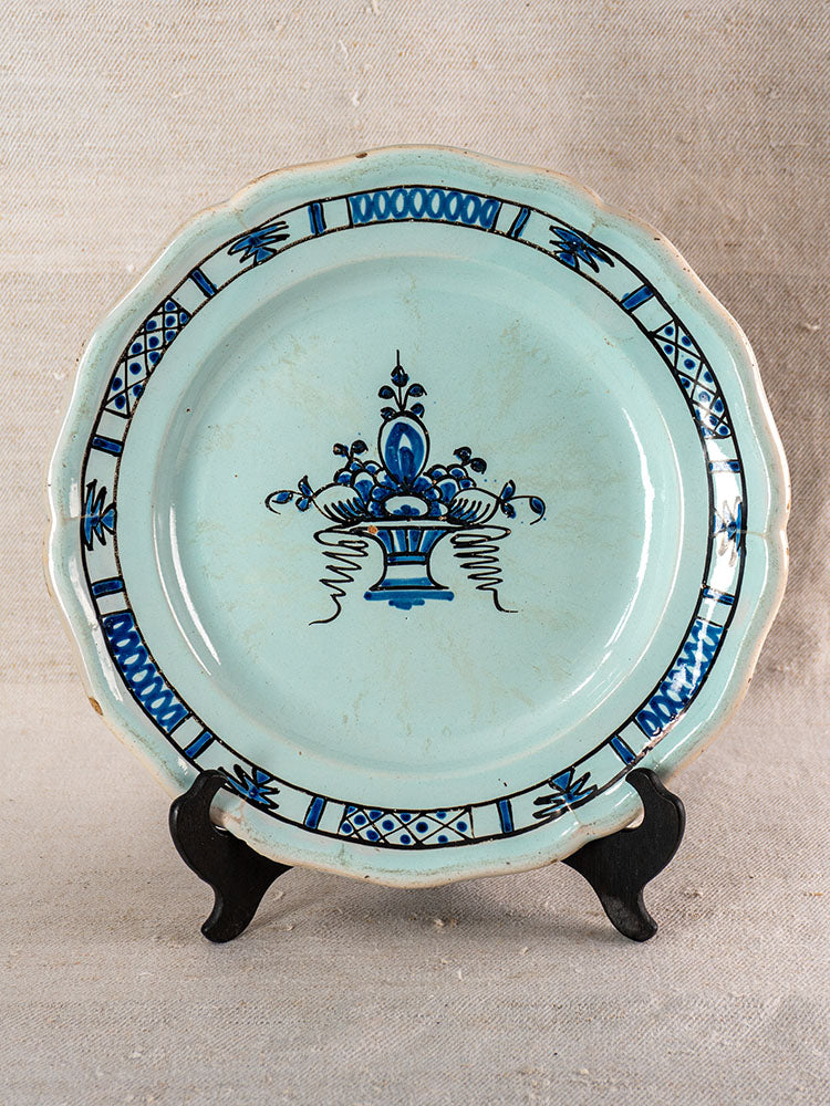 Antique French faience plate with light blue glaze, ornate blue floral center, scalloped rim on black stand, beige background.