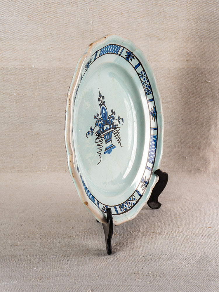 Antique French faience plate with central blue floral and geometric rim, on black stand, beige textured background.