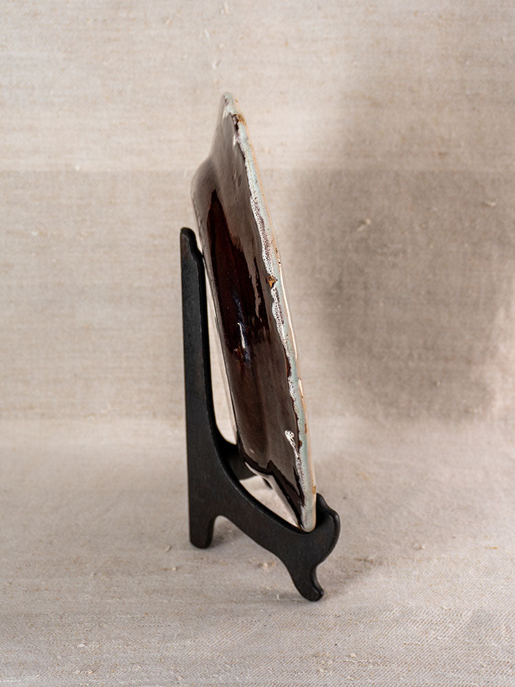 19th-century French faience Cul Noir plate with dark glossy glaze on black stand, neutral textured fabric backdrop.