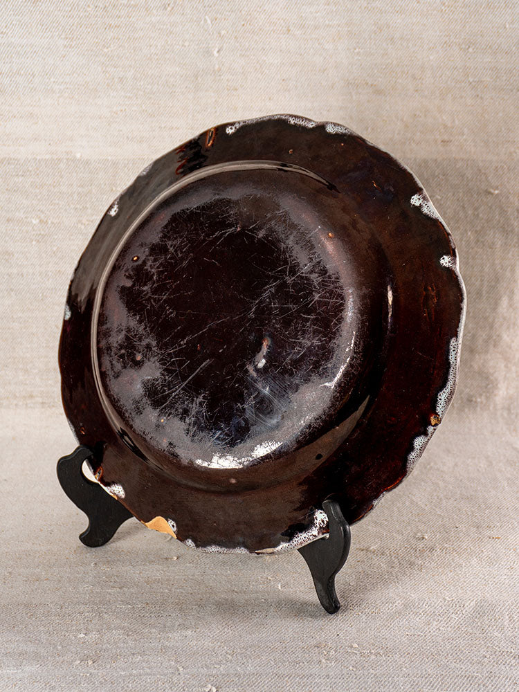 Antique French faience plate with dark wavy rim, visible scuffs and chips, on black stand against neutral fabric background.