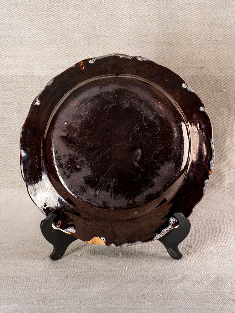 19th-century French faience Cul Noir plate with irregular rim and mottled brown glaze, upright on beige fabric. Rouen style inspired.
