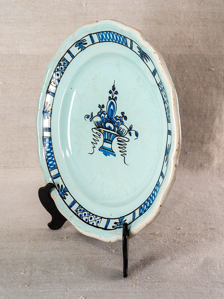 19th century French faience oval plate with blue glaze, floral basket motif, and decorative border on black stand.