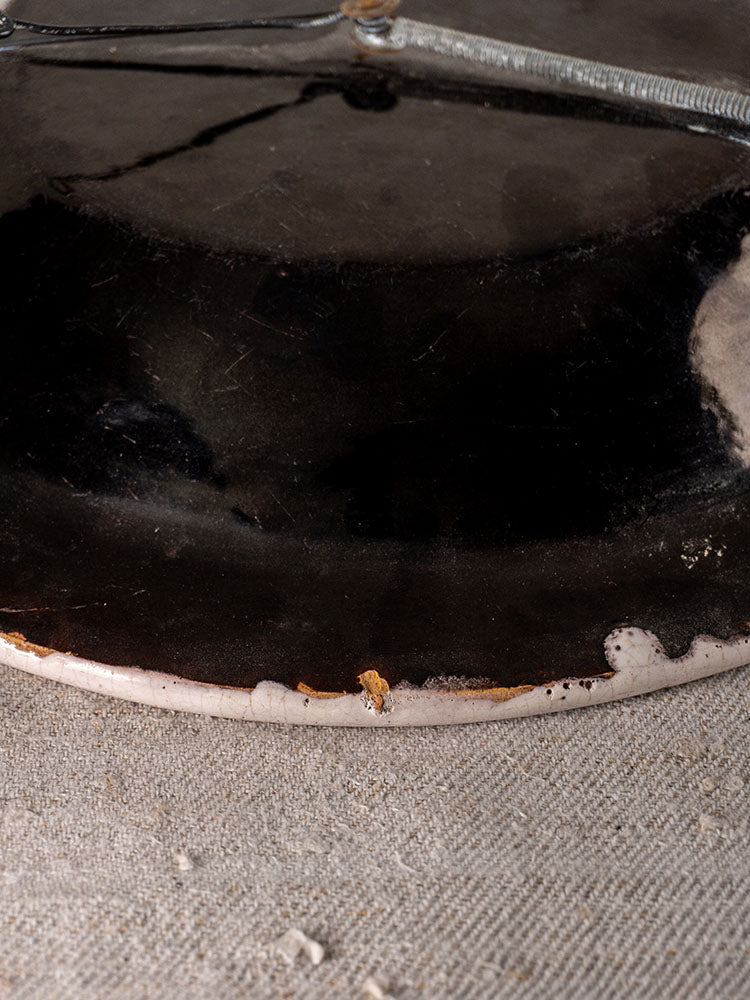 Antique French Cul Noir faience charger with chipped glaze, exposed ceramic edge, on textured beige fabric.