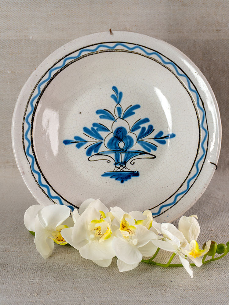 Antique French faience Cul Noir charger with Panier Fleuri design, displayed upright with white orchid on neutral fabric.
