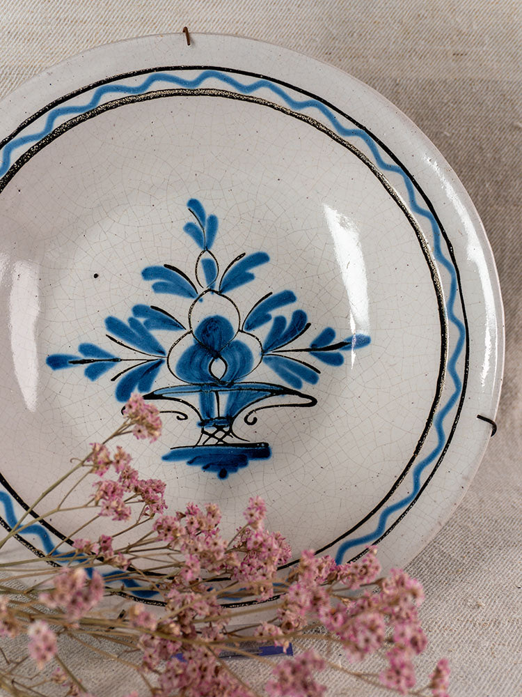 Antique French Cul Noir faience charger with blue floral decor, black rim, on beige fabric with pink dried flowers.