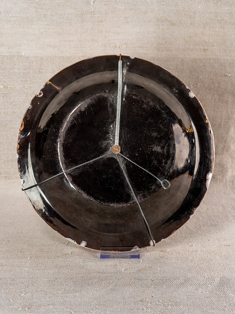 Antique French cul noir faience charger with panier fleuri, rim chips stapled, upright on clear stand against beige fabric.