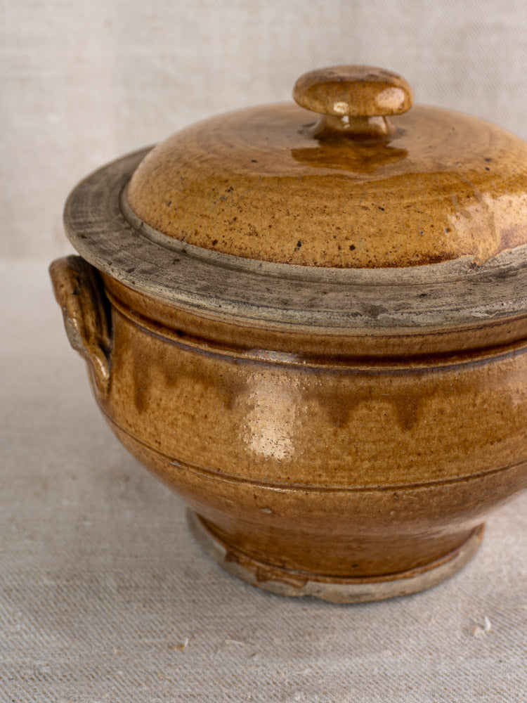 Antique French lidded stoneware pot with honey glazed grès finish and side handle on textured fabric.
