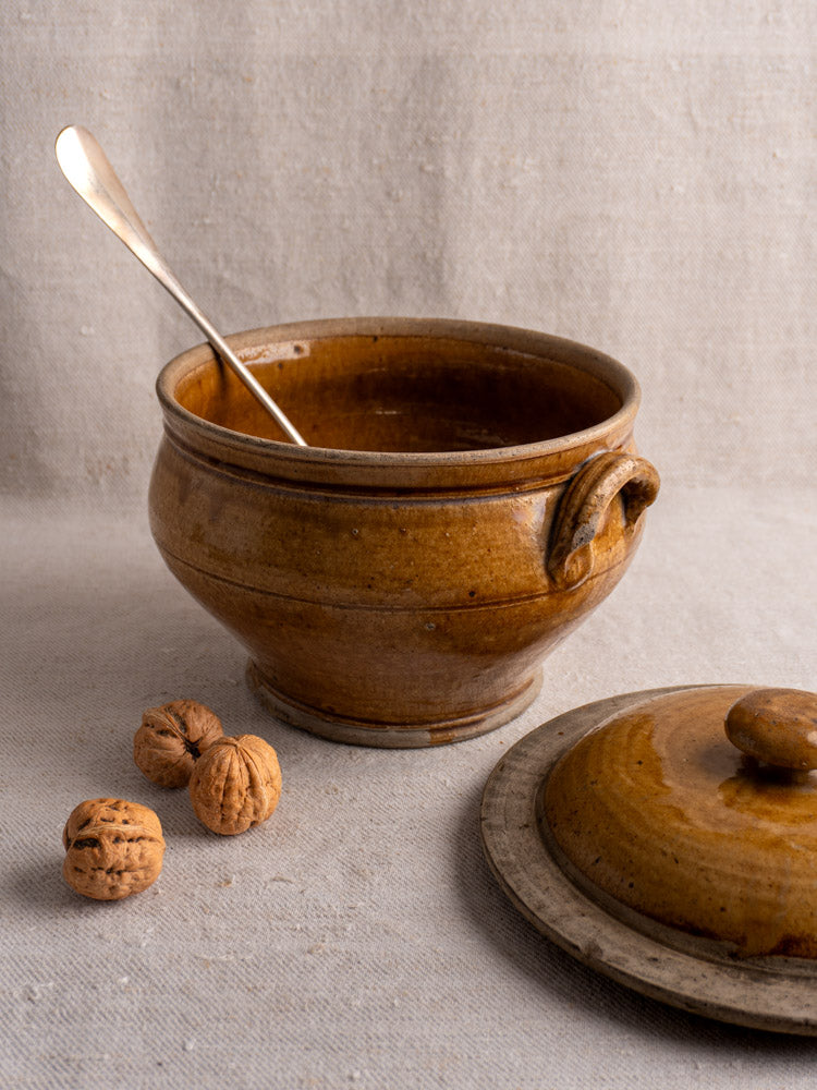 Antique French honey-glazed stoneware pot with lid, metal spoon inside, walnuts beside on fabric.