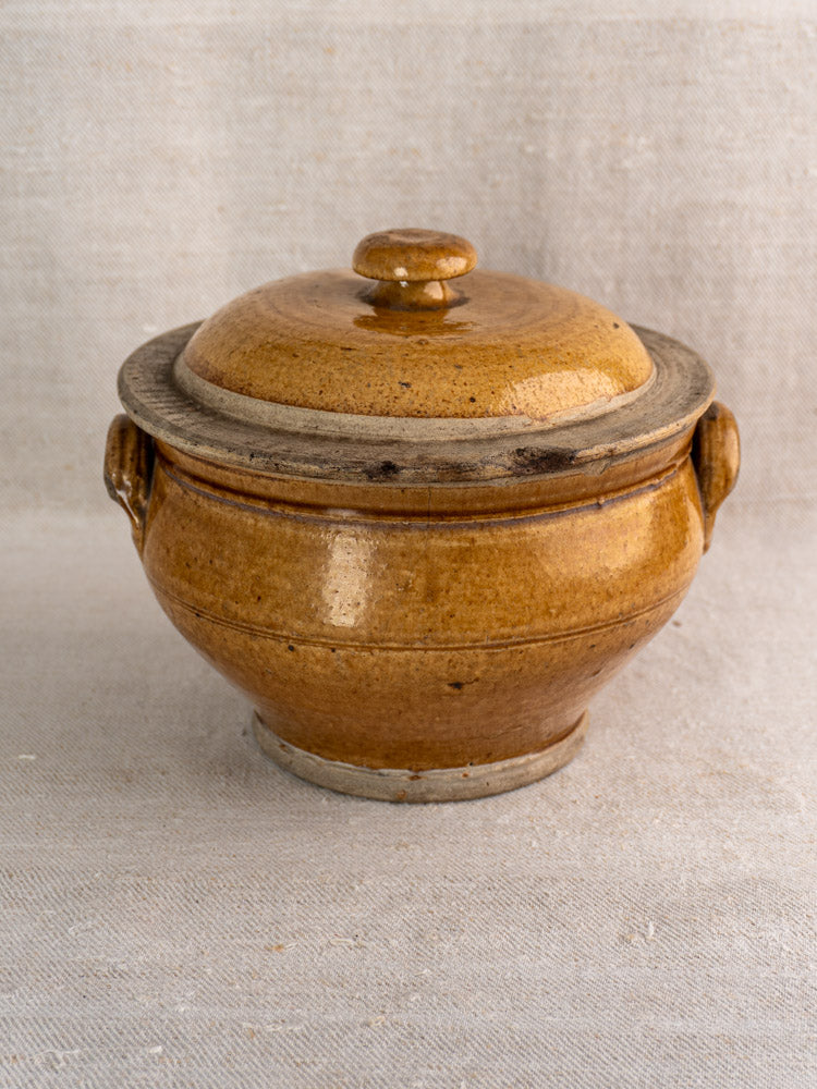 Antique French honey-glazed stoneware pot with lid and side handles on light fabric background.