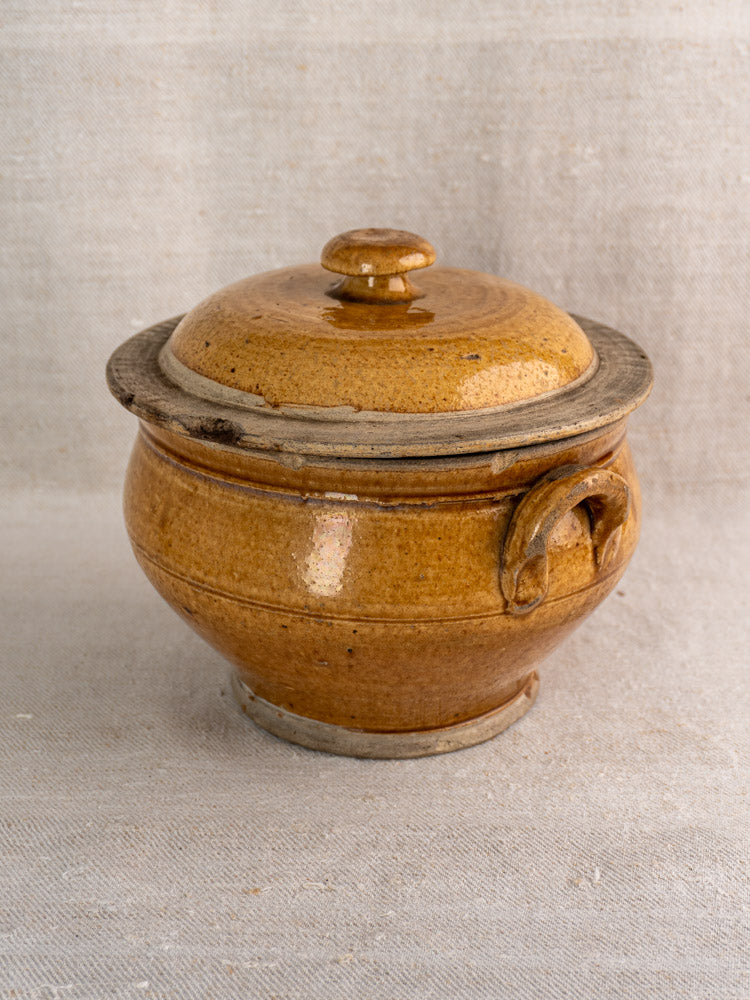 Antique French lidded stoneware pot with honey glaze, rustic rim, on textured beige fabric.