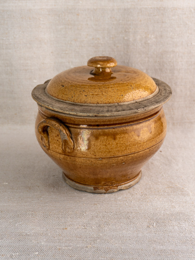 Antique French lidded stoneware pot with handles, honey brown glaze, on light textured fabric.