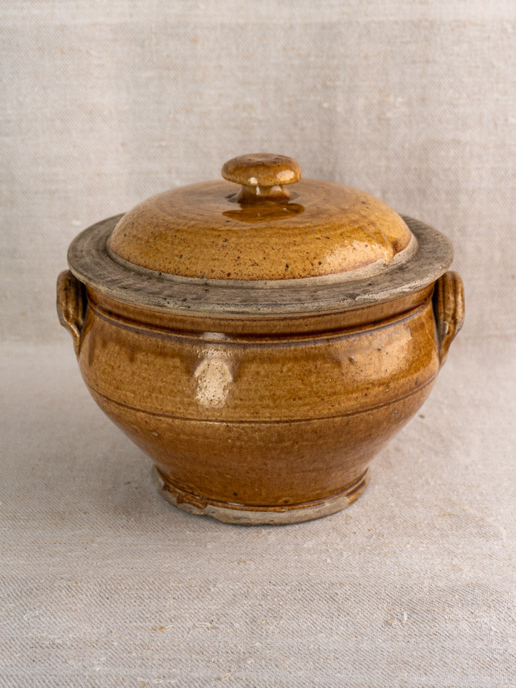 Antique French 19th-century stoneware pot with lid and side handles, honey glaze, on fabric.