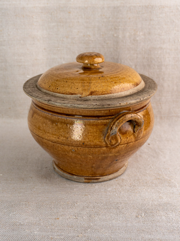 Antique French lidded stoneware pot in glossy honey brown grès with curved handle on beige textile.