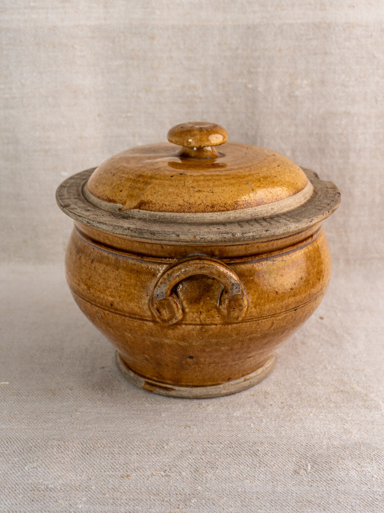 Antique French lidded stoneware pot with rich honey glaze on light textured fabric background.