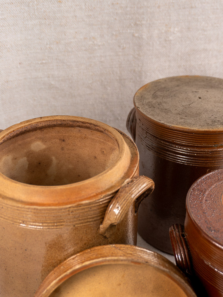 Set of three antique French stoneware confit pots with lids displayed on neutral textured fabric.
