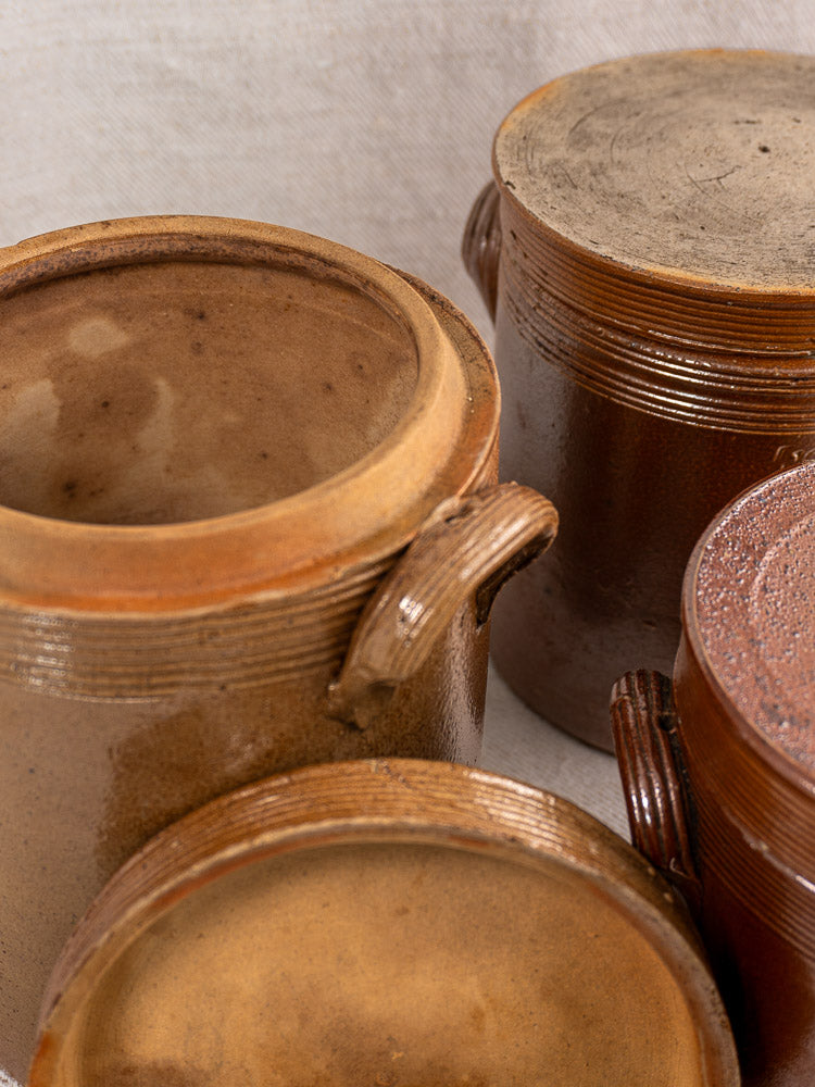 Three antique French stoneware confit pots with brown glaze and lids displayed on neutral fabric.