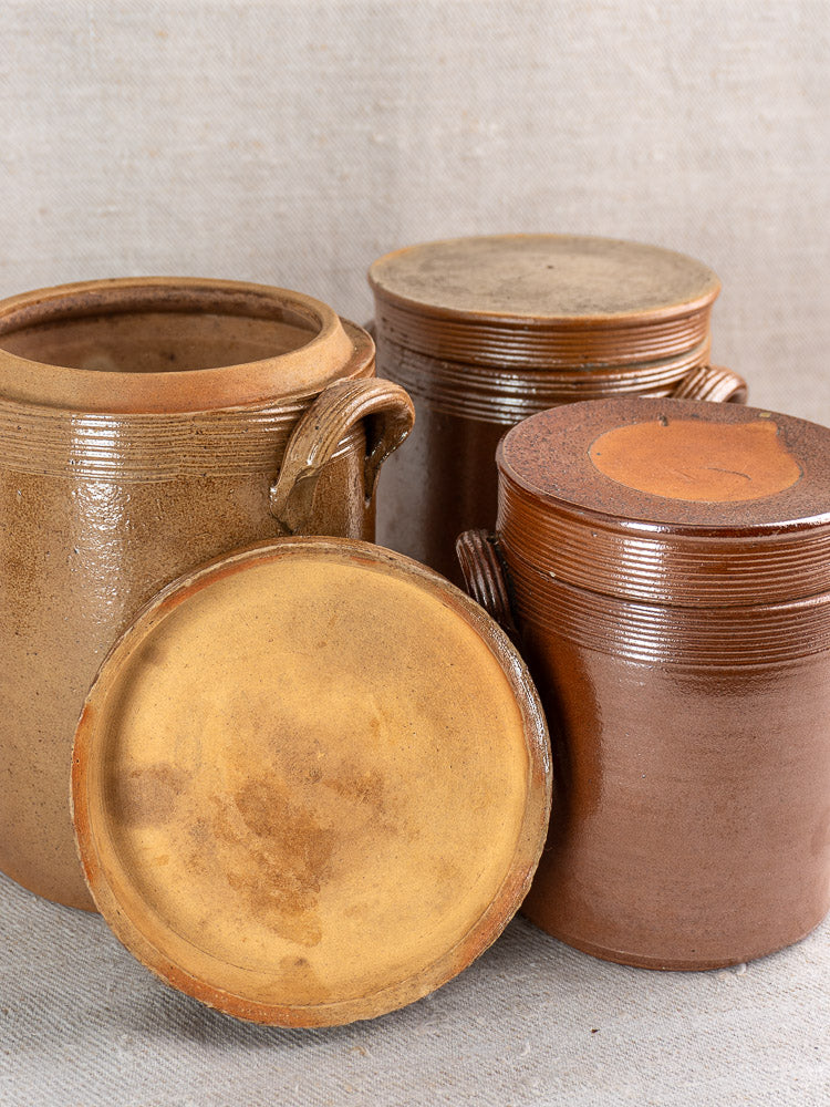 Set of three antique French stoneware confit pots with lids, one open, on textured beige fabric.