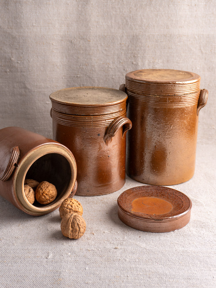 Set of three antique French stoneware confit pots with lids, one tipped with walnuts, on a textured surface.