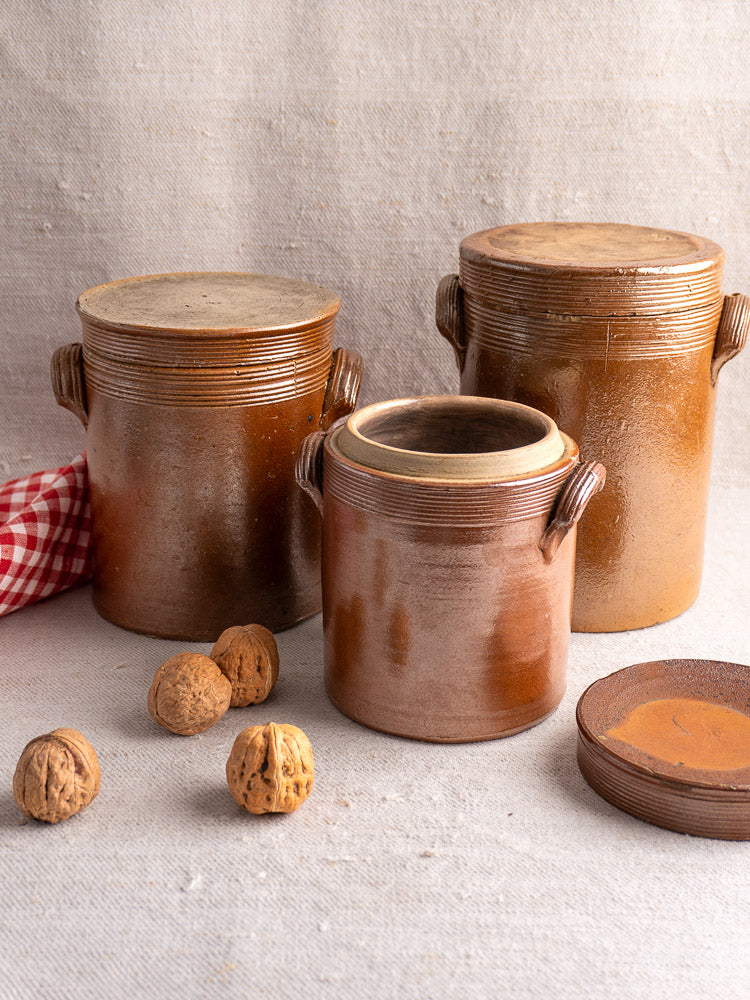 Set of three antique French stoneware confit pots with lids, walnuts, dish, and checked cloth on rustic kitchen surface.