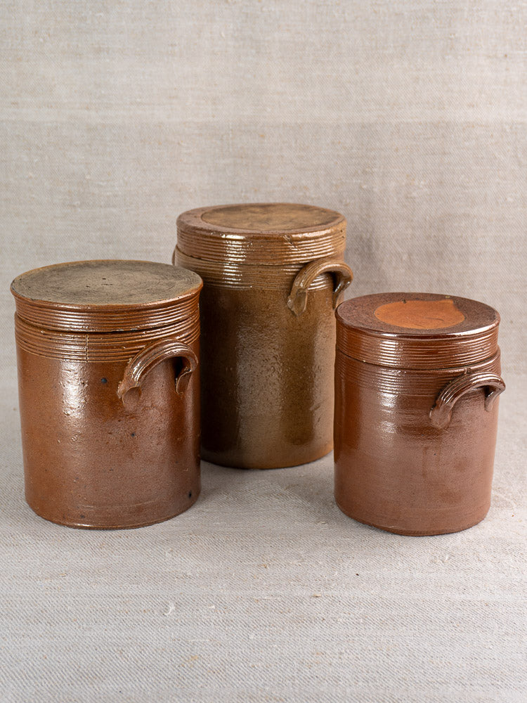 Set of three antique French stoneware confit pots with lids and side handles on neutral fabric background.