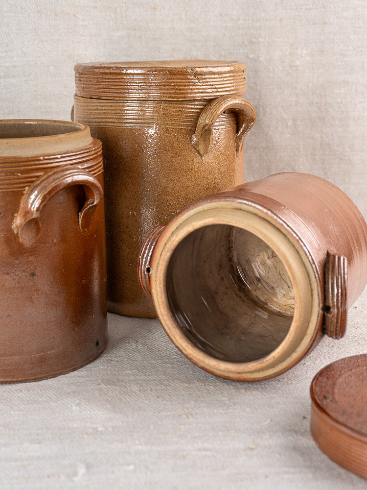 Set of three antique French stoneware confit pots, one tipped to reveal glazed interior, on beige fabric.