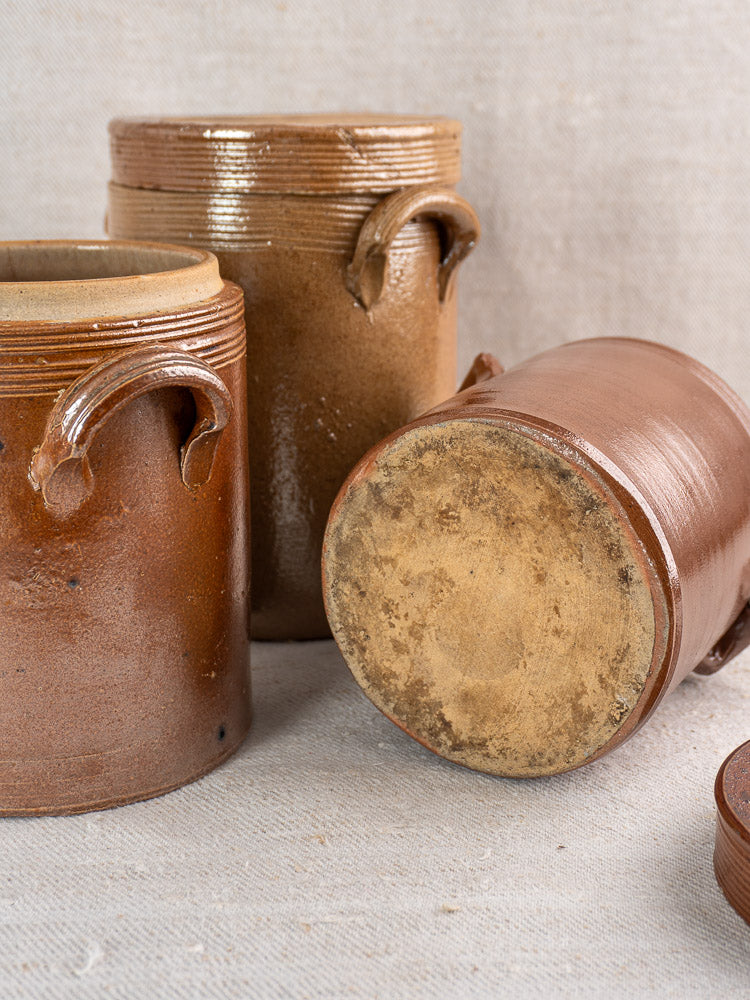 Set of three antique French stoneware confit pots with handles, two upright, one on textured fabric.